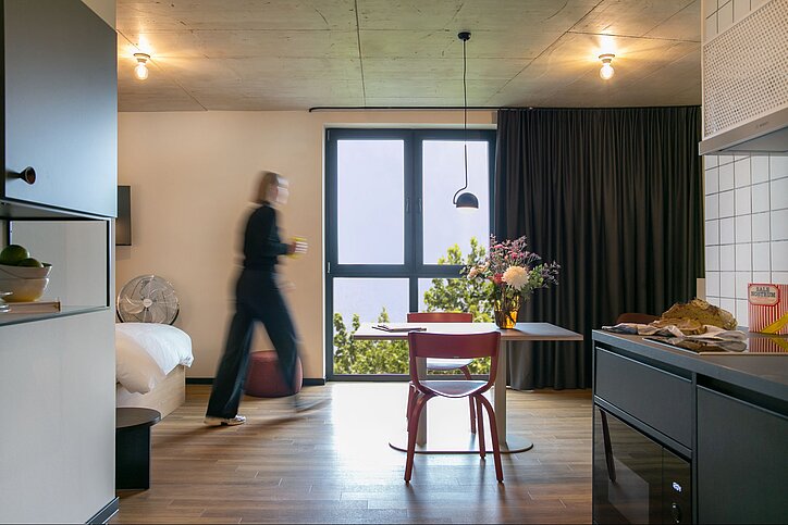 Stayery hat jetzt auch in der Ruhrstadt Bochum eine Adresse. Das Gebäude hatte Catella 2023 gekauft. © Stayery Apartment mit Fenster, Tisch, Küchenzeile und einer Person, die durchläuft